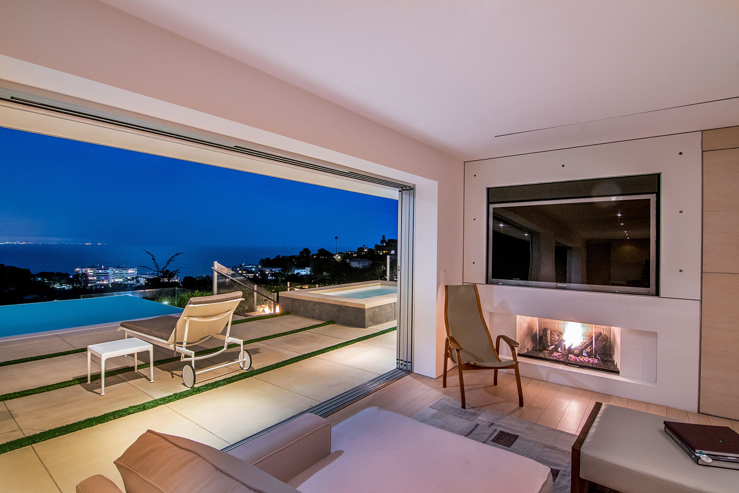 Contemporary living room with fireplace opening to an infinity pool and panoramic Pacific Ocean views at twilight.